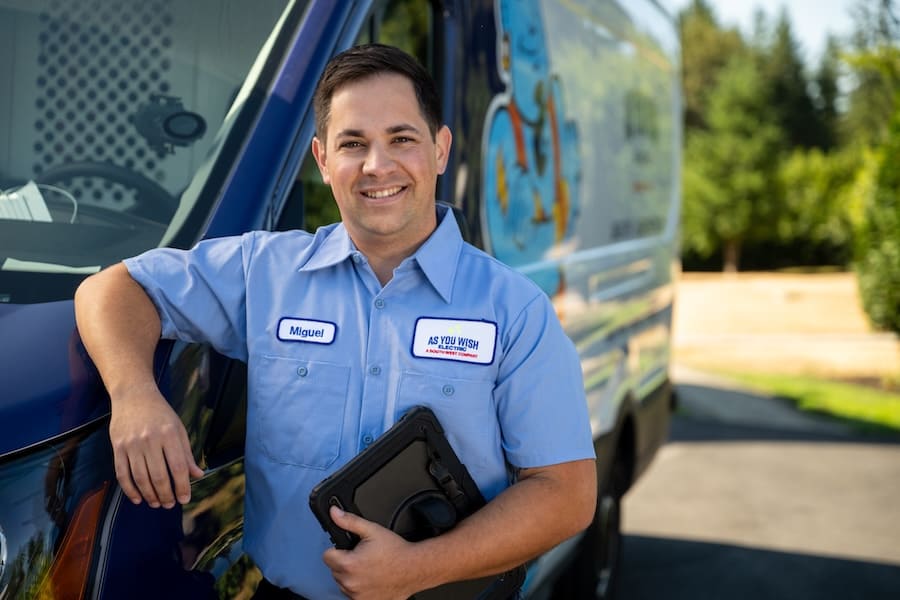 Maintenance technician Miguel poses beside a blue company van, holding a black tablet case and wearing a light blue uniform with a name patch and a company patch reading 'AS YOU WISH'.